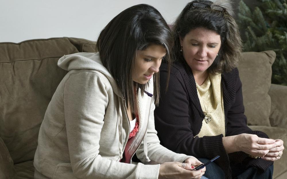A woman and her teenage daughter sit on the couch discussing something that the teen is reading on her mobile phone.