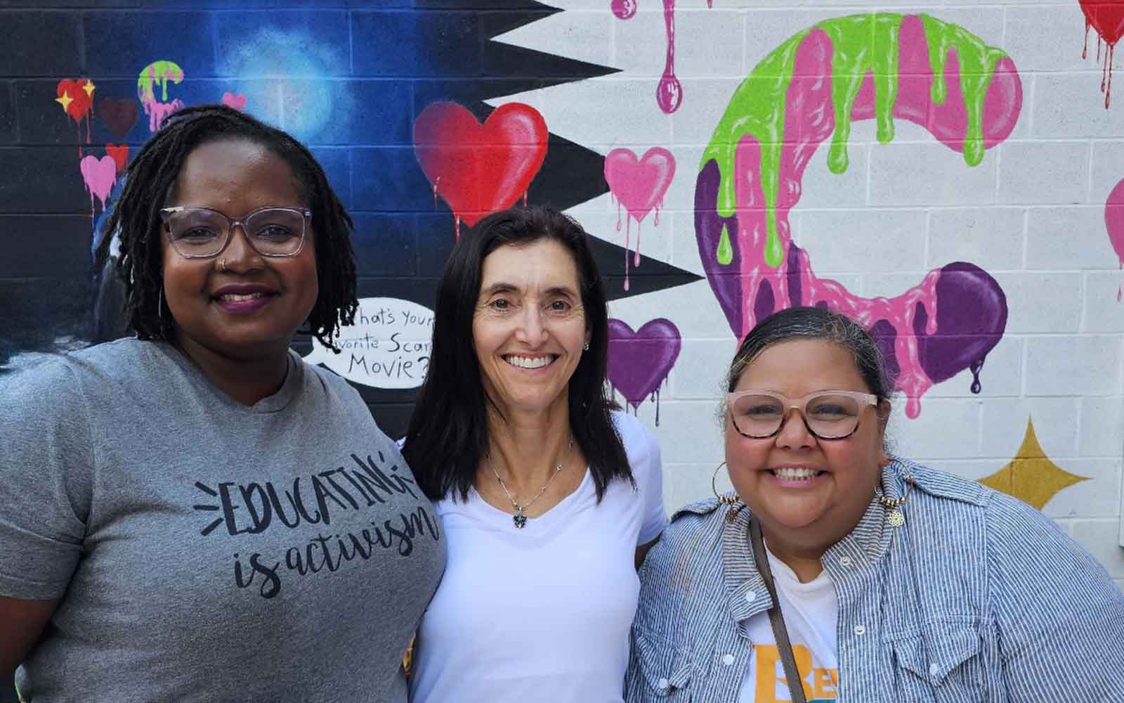 photo of three smiling women standing in front of a mural